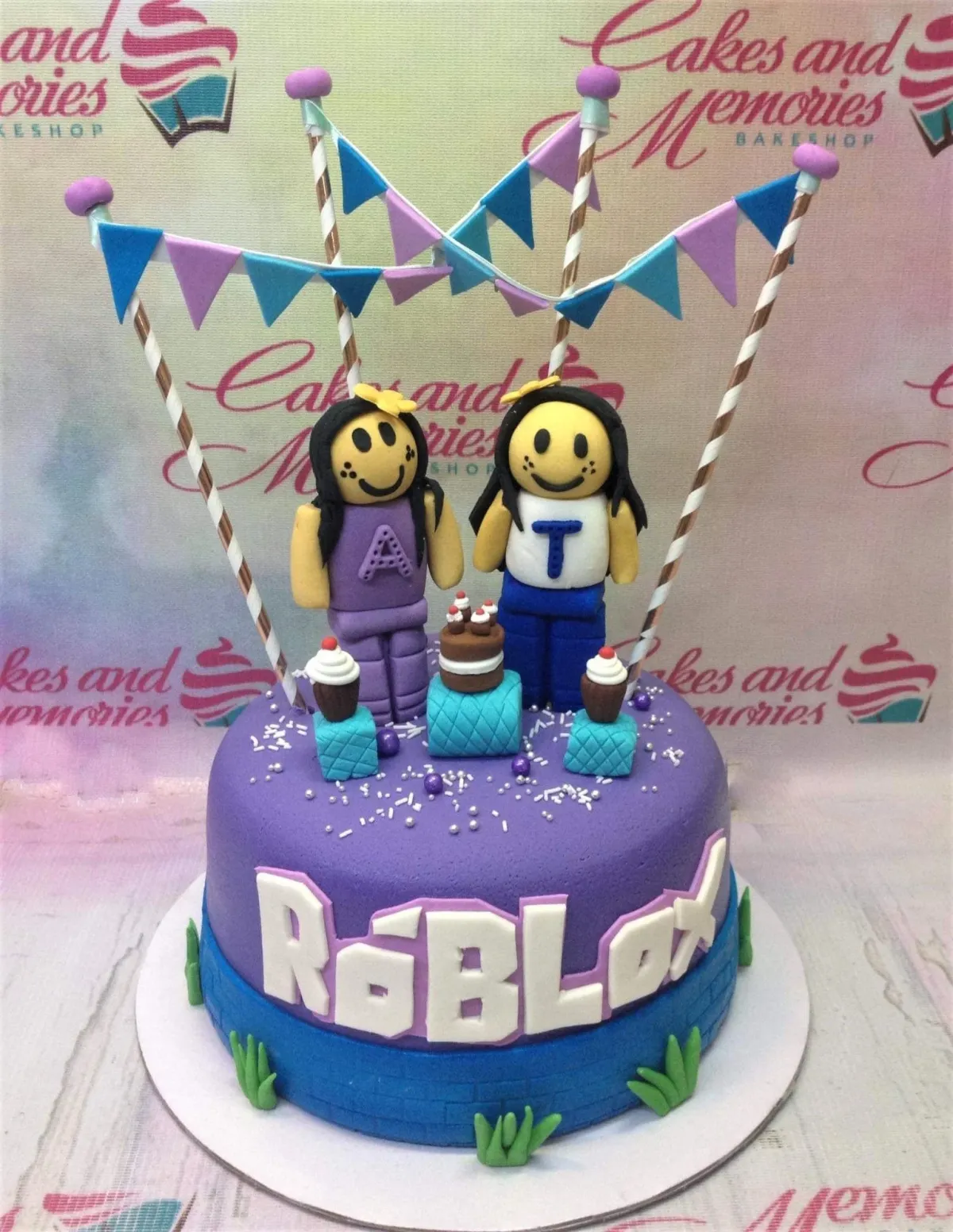 A purple and blue fondant cake themed around Roblox, featuring two girl figures, bunting flags, and a brick-textured base. A purple and blue fondant cake themed around Roblox, featuring two girl figures, bunting flags, and a brick-textured base.