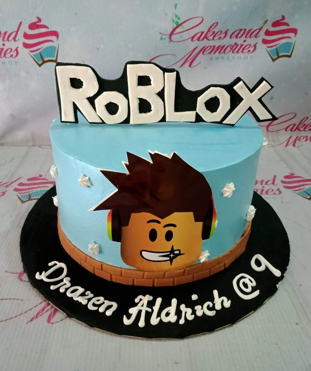 A light blue 1-tier cake themed around Roblox, featuring a Roblox character printout, a Roblox logo banner, and a brick-patterned base border. A light blue 1-tier cake themed around Roblox, featuring a Roblox character printout, a Roblox logo banner, and a brick-patterned base border.