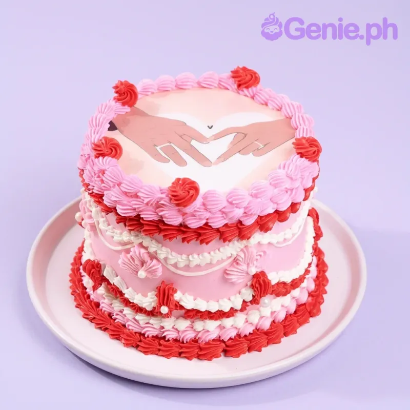 A pink and white 1-tier cake with red accents, Romantic Vintage themed, featuring a hand-heart printout and ruffled icing.