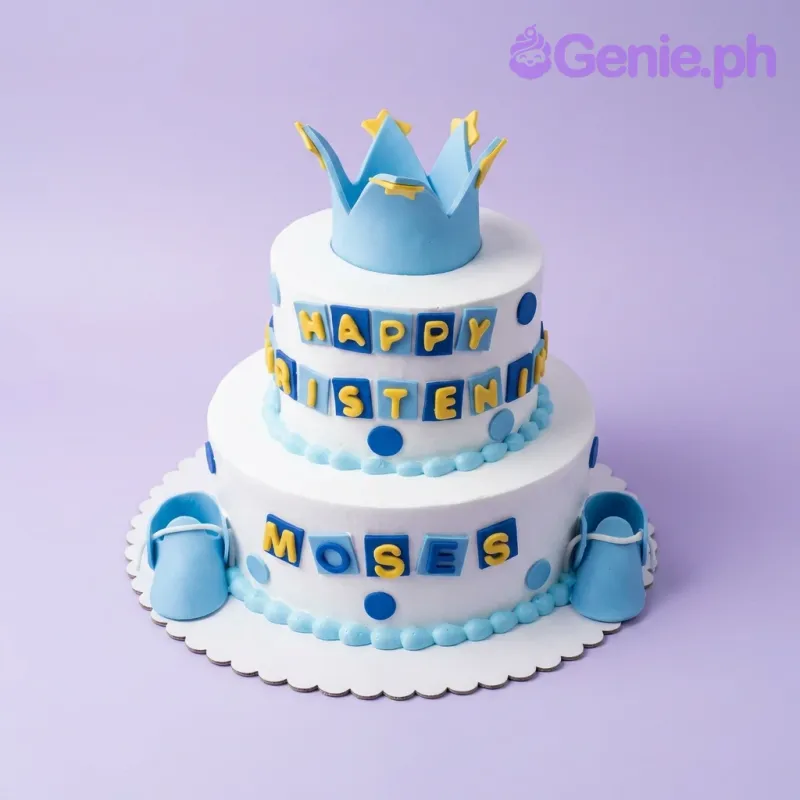 Two-tier white fondant cake with a blue crown topper, blue baby shoes, and blue and yellow gumpaste letter tiles. Two-tier white fondant cake with a blue crown topper, blue baby shoes, and blue and yellow gumpaste letter tiles.