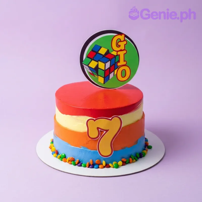 A red, yellow, orange, and blue 1 Tier cake themed around Rubik's Cube, featuring a circular Rubik's Cube topper with 'Gio' and a large yellow number 7, with colorful chocolate candies around the base. A red, yellow, orange, and blue 1 Tier cake themed around Rubik's Cube, featuring a circular Rubik's Cube topper with 'Gio' and a large yellow number 7, with colorful chocolate candies around the base.