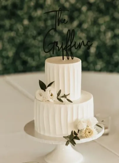 White 2-tier rustic wedding cake with soft icing, vertical ribbed textures, a black cardstock name topper, and white fondant flowers White 2-tier rustic wedding cake with soft icing, vertical ribbed textures, a black cardstock name topper, and white fondant flowers