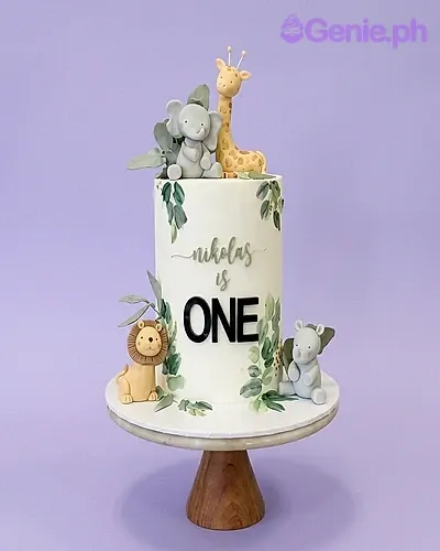 A white single tier cake themed around safari animals, featuring fondant lion, giraffe, and elephant figures and green tropical leaves. A white single tier cake themed around safari animals, featuring fondant lion, giraffe, and elephant figures and green tropical leaves.