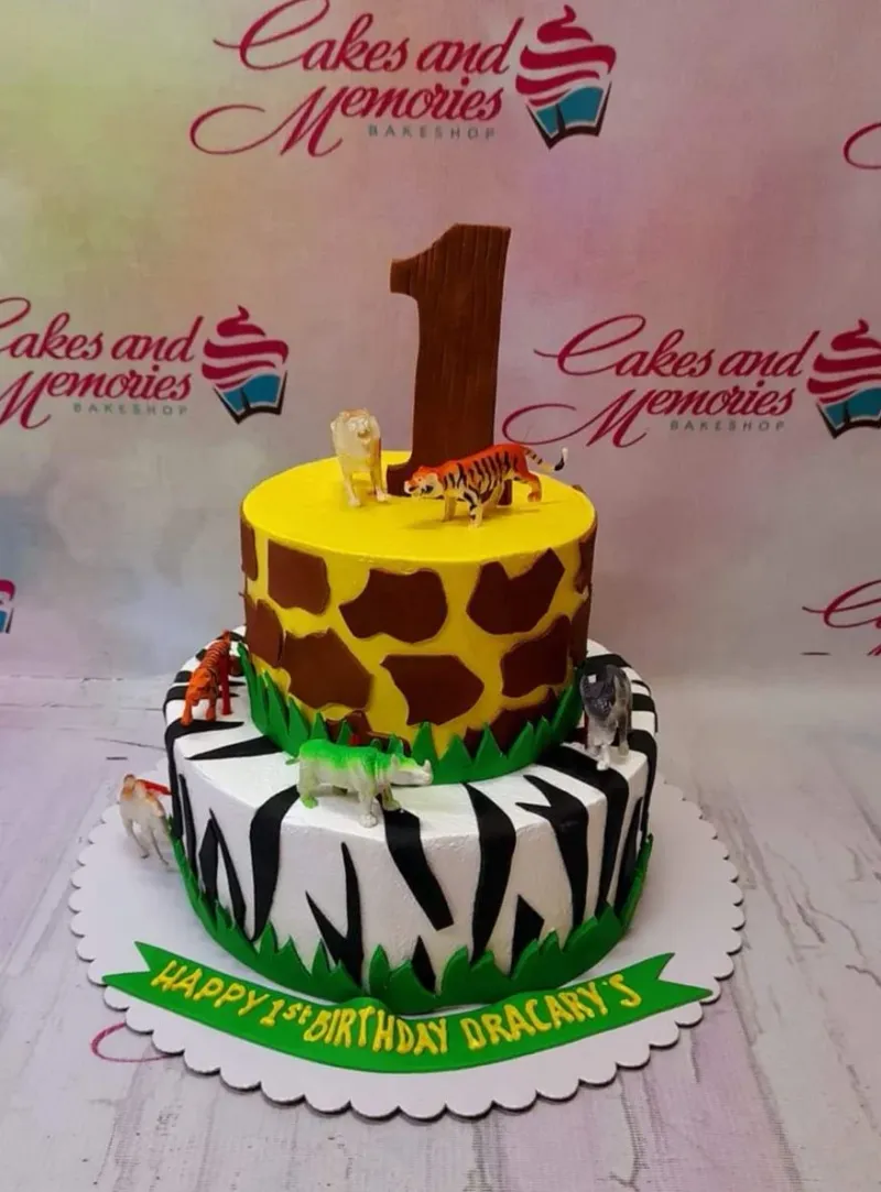 A yellow and white two-tier Safari Animal cake, featuring a brown number one, plastic safari animals, giraffe spots, and zebra stripes. A yellow and white two-tier Safari Animal cake, featuring a brown number one, plastic safari animals, giraffe spots, and zebra stripes.