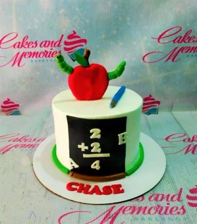 A white 1-tier school themed cake, featuring a large red apple with a green worm, a blue pencil, and a black chalkboard. A white 1-tier school themed cake, featuring a large red apple with a green worm, a blue pencil, and a black chalkboard.