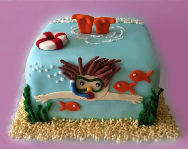 Square light blue fondant cake with a 3D scuba diver figure, orange fish, and a lifebuoy on top, surrounded by puffed rice sand. Square light blue fondant cake with a 3D scuba diver figure, orange fish, and a lifebuoy on top, surrounded by puffed rice sand.