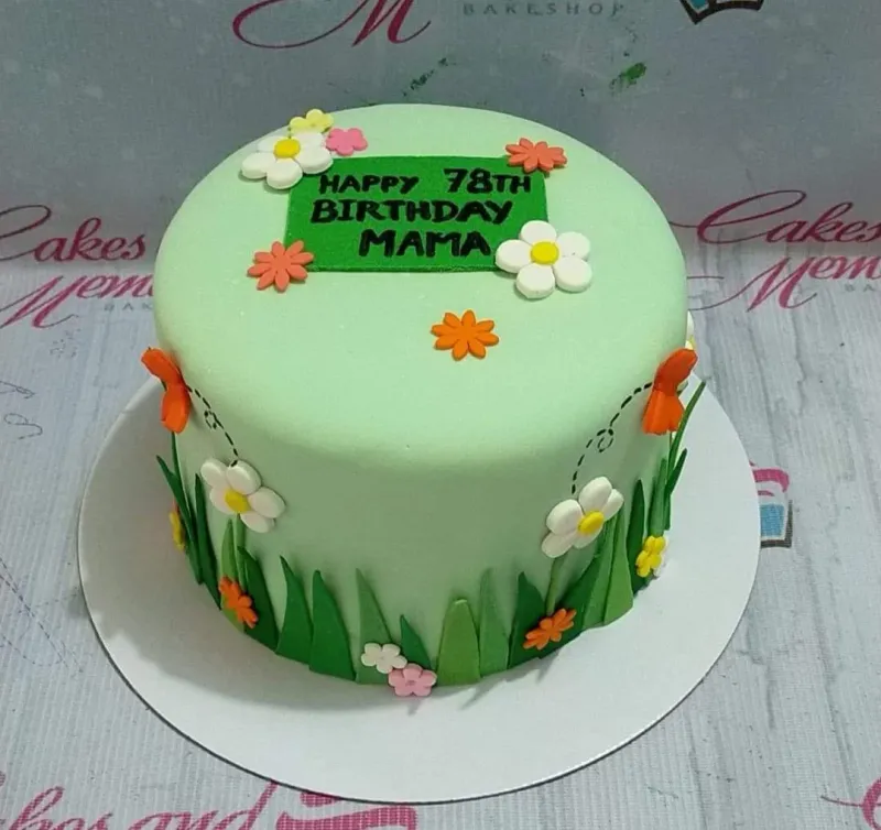 A light green 1-tier fondant cake for a senior birthday, featuring edible flowers, grass details, and a green message plaque. A light green 1-tier fondant cake for a senior birthday, featuring edible flowers, grass details, and a green message plaque.