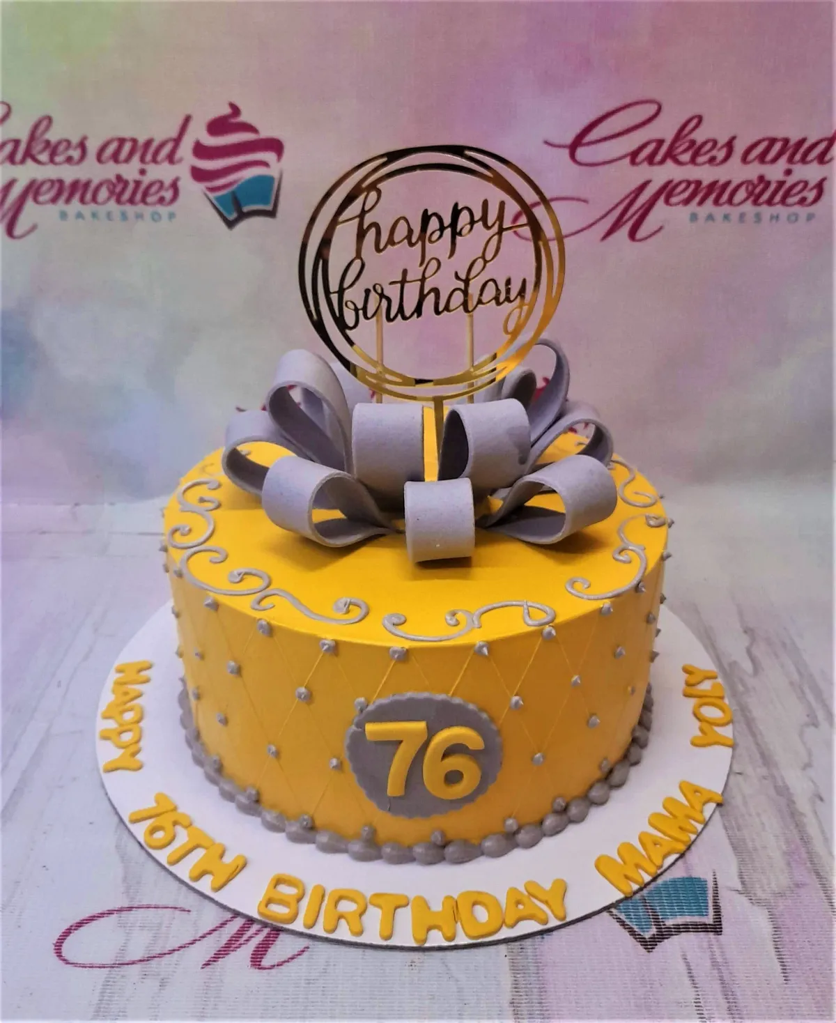 Yellow 1-tier birthday cake with a large grey fondant bow, gold Happy Birthday topper, and grey quilted pattern with silver beads. Yellow 1-tier birthday cake with a large grey fondant bow, gold Happy Birthday topper, and grey quilted pattern with silver beads.