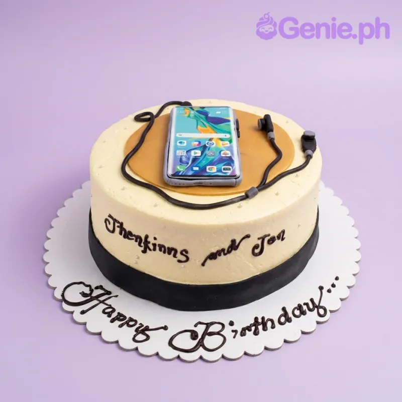 A round cream-colored cake featuring a smartphone printout and fondant headphones on top, with a black base band and birthday message. A round cream-colored cake featuring a smartphone printout and fondant headphones on top, with a black base band and birthday message.