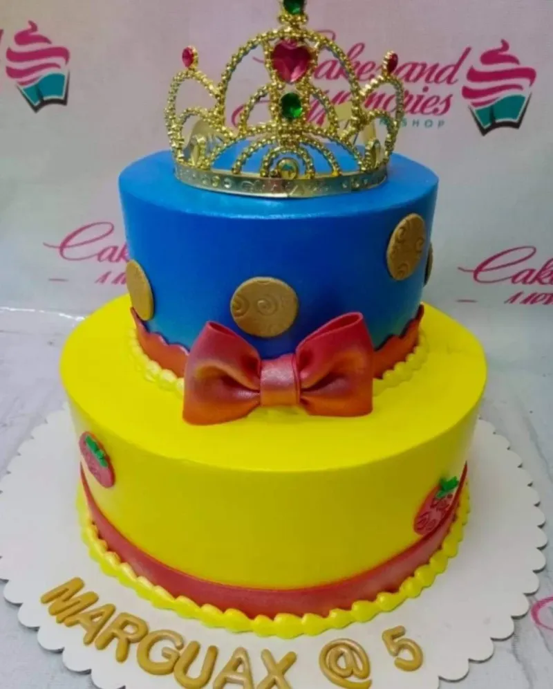 Two-tier Snow White themed cake with a blue top tier, yellow bottom tier, red bow, and a gold rhinestone crown topper. Two-tier Snow White themed cake with a blue top tier, yellow bottom tier, red bow, and a gold rhinestone crown topper.