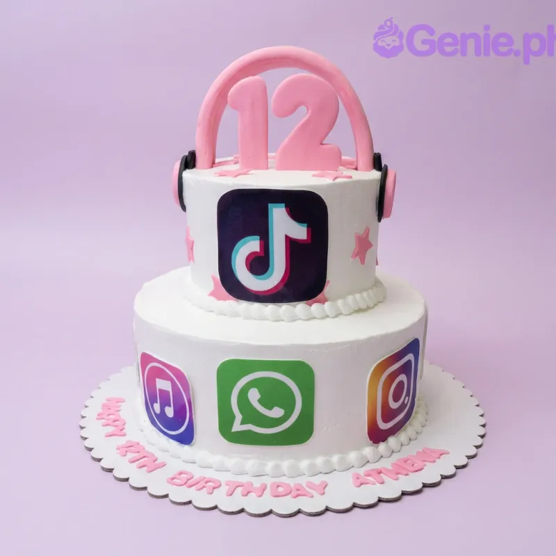 A white two-tier cake themed around Social Media, featuring pink headphones, a number 12 topper, and app icons like TikTok, Instagram, and WhatsApp. A white two-tier cake themed around Social Media, featuring pink headphones, a number 12 topper, and app icons like TikTok, Instagram, and WhatsApp.