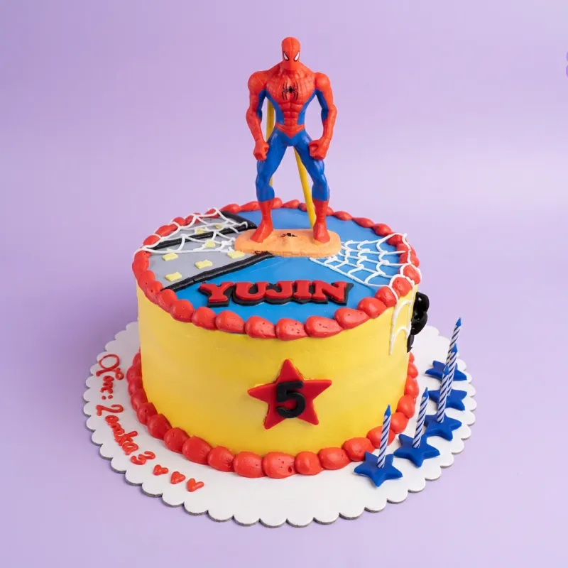 A yellow, blue, red, and white 1-tier Spiderman cake with a 3D Spiderman figure, a red star, white spiderwebs, and red icing borders. A yellow, blue, red, and white 1-tier Spiderman cake with a 3D Spiderman figure, a red star, white spiderwebs, and red icing borders.