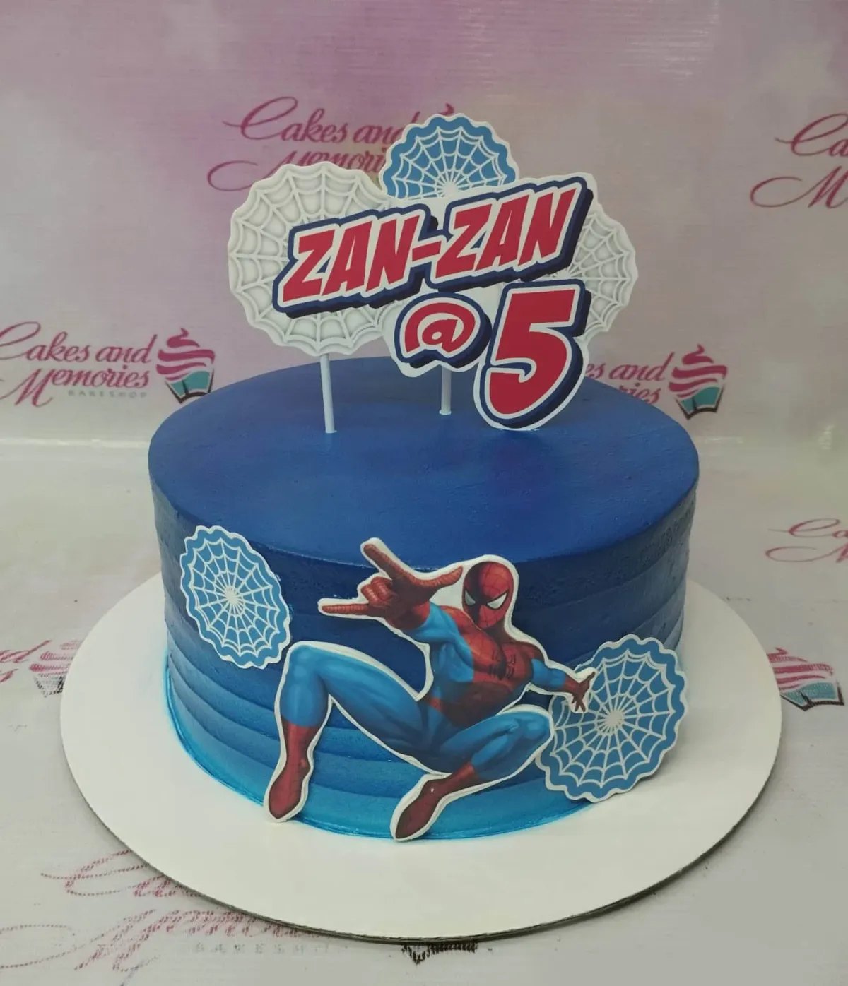 A blue 1-tier cake themed around Spiderman, featuring a Spiderman character and a personalized name and age topper, with white spider web accents. A blue 1-tier cake themed around Spiderman, featuring a Spiderman character and a personalized name and age topper, with white spider web accents.