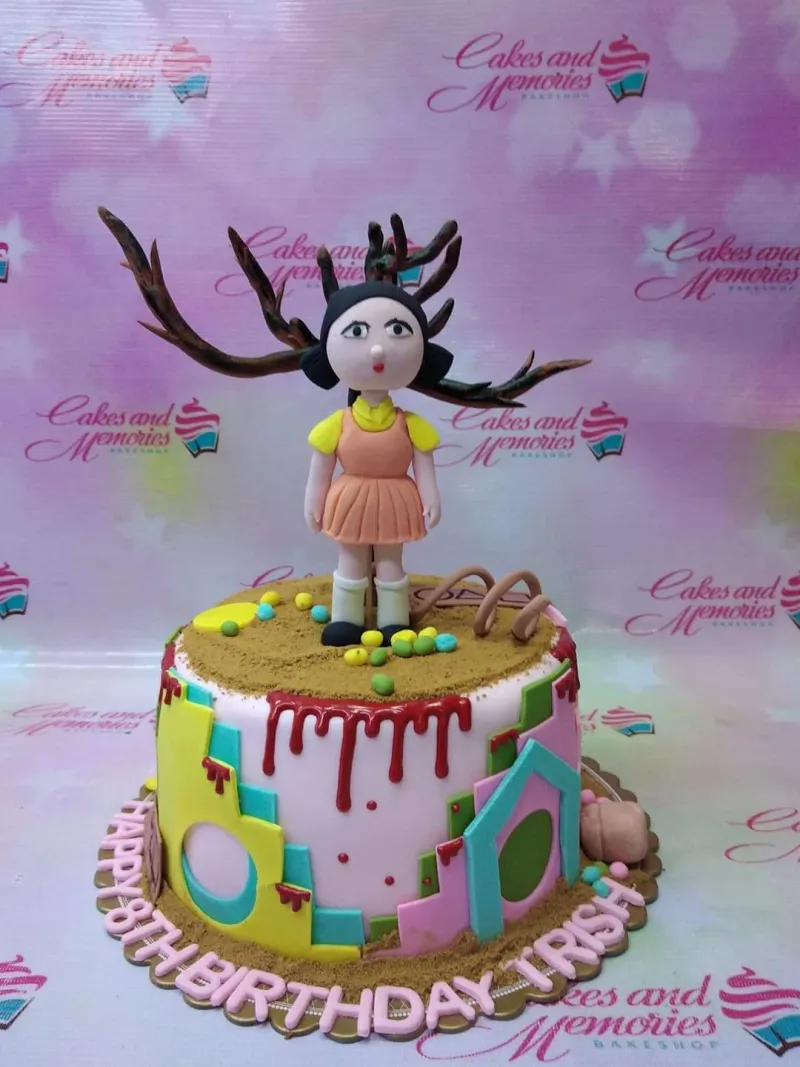 A pink 1 Tier Fondant cake themed around Squid Game, featuring an iconic giant doll, geometric panels, and a red drip. A pink 1 Tier Fondant cake themed around Squid Game, featuring an iconic giant doll, geometric panels, and a red drip.