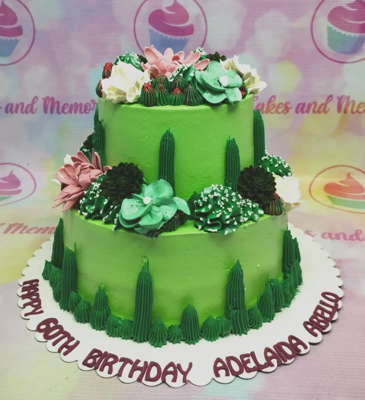 A green two-tier cake themed around a Succulent Garden, featuring pink, white, and teal icing flowers and dark green cactus piping. A green two-tier cake themed around a Succulent Garden, featuring pink, white, and teal icing flowers and dark green cactus piping.