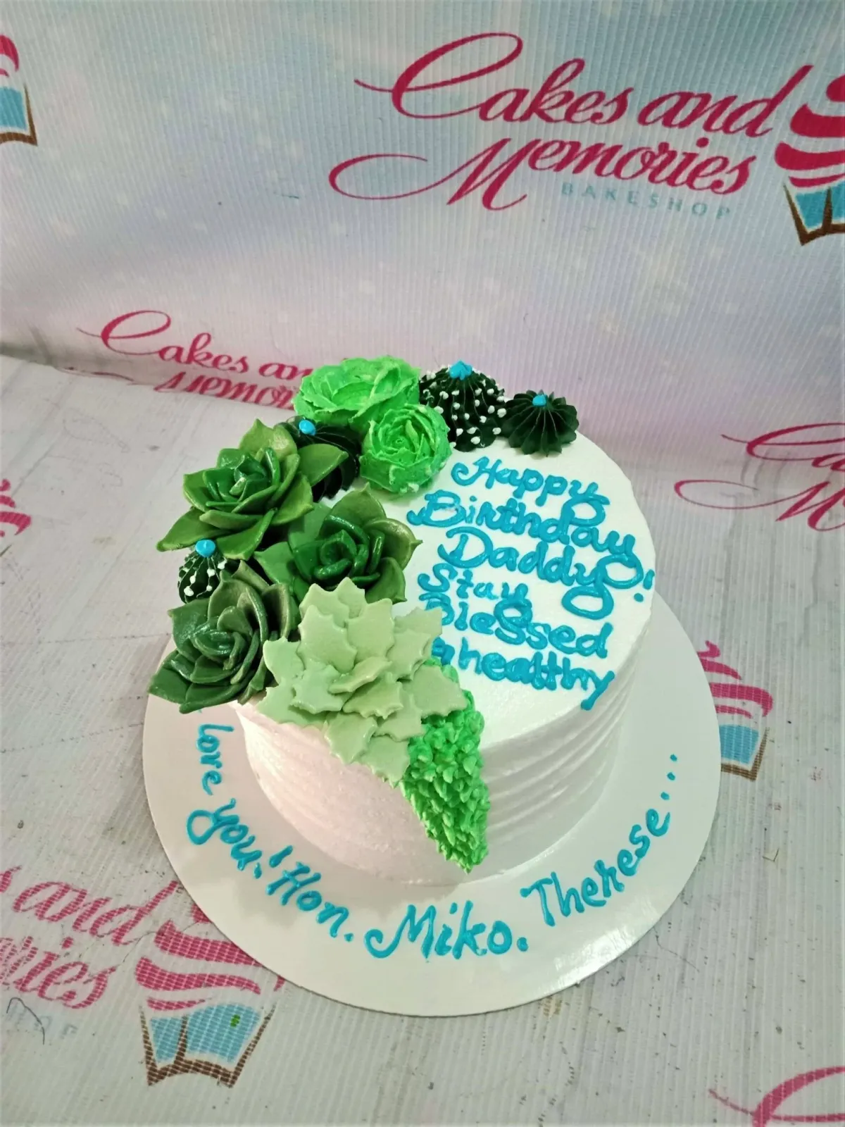 A white and green round 1-tier cake themed as a Succulent Garden, featuring piped icing succulents and cacti, and blue text. A white and green round 1-tier cake themed as a Succulent Garden, featuring piped icing succulents and cacti, and blue text.