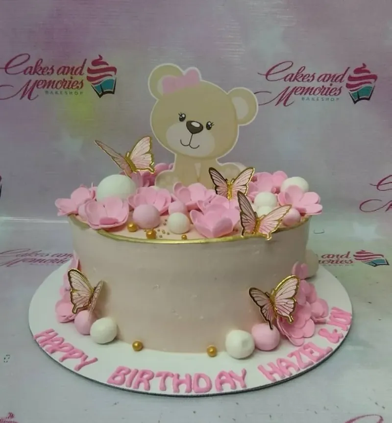 Pink teddy bear birthday cake with pink fondant flowers, gold butterflies, and white spheres on a white base board. Pink teddy bear birthday cake with pink fondant flowers, gold butterflies, and white spheres on a white base board.