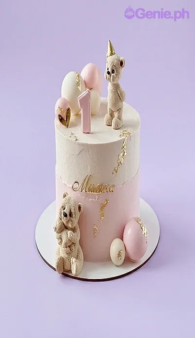 A pink and white 1-tier Teddy Bear cake, featuring two hand-sculpted teddy bears, a pink number one, and gold leaf accents. A pink and white 1-tier Teddy Bear cake, featuring two hand-sculpted teddy bears, a pink number one, and gold leaf accents.