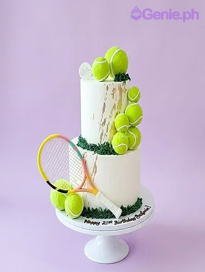 White 2-tier tennis themed cake with green grass icing, gold brush strokes, and multiple tennis ball toppers with a racket. White 2-tier tennis themed cake with green grass icing, gold brush strokes, and multiple tennis ball toppers with a racket.
