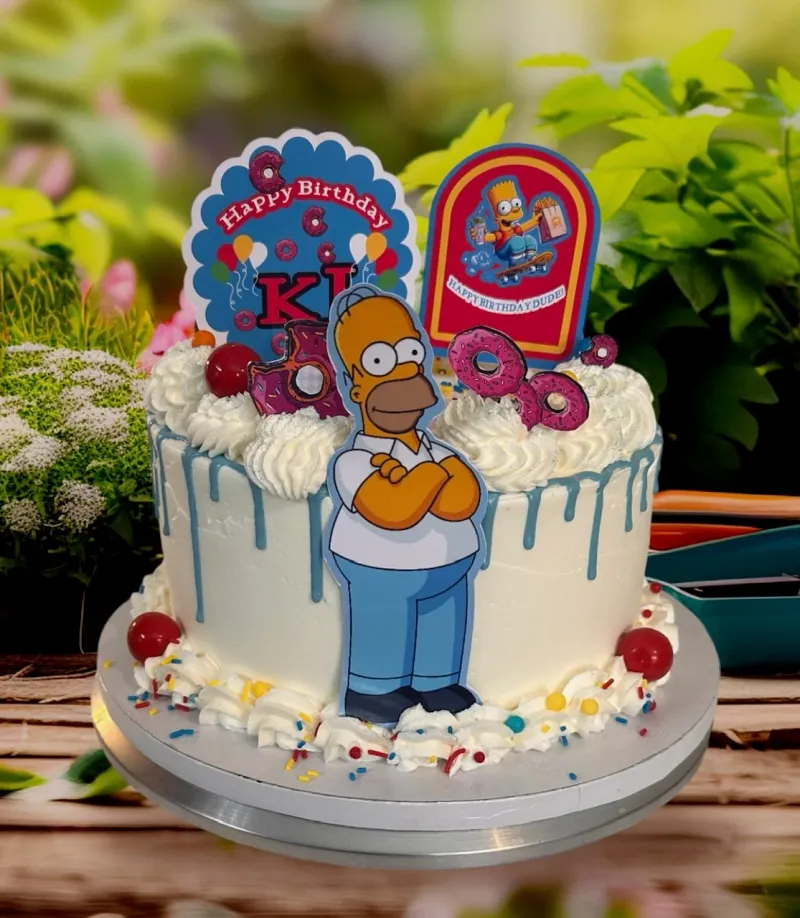 A white 1-tier cake themed around The Simpsons, featuring a Homer Simpson printout, blue icing drip, and donut decorations. A white 1-tier cake themed around The Simpsons, featuring a Homer Simpson printout, blue icing drip, and donut decorations.