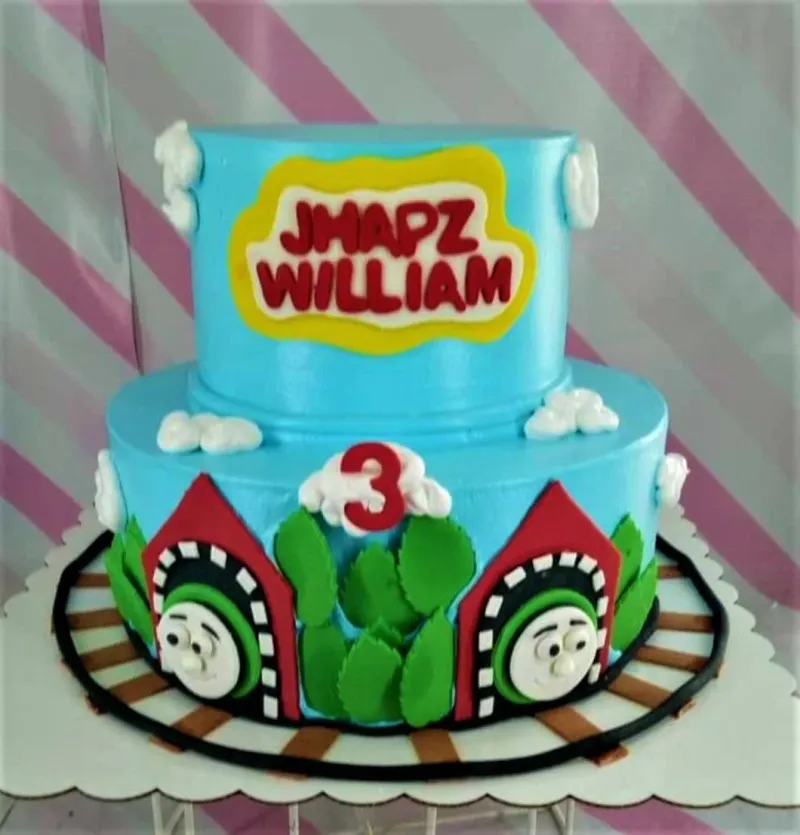 Two-tier light blue Thomas the Tank Engine themed birthday cake with fondant train tunnels, green bushes, and white clouds. Two-tier light blue Thomas the Tank Engine themed birthday cake with fondant train tunnels, green bushes, and white clouds.