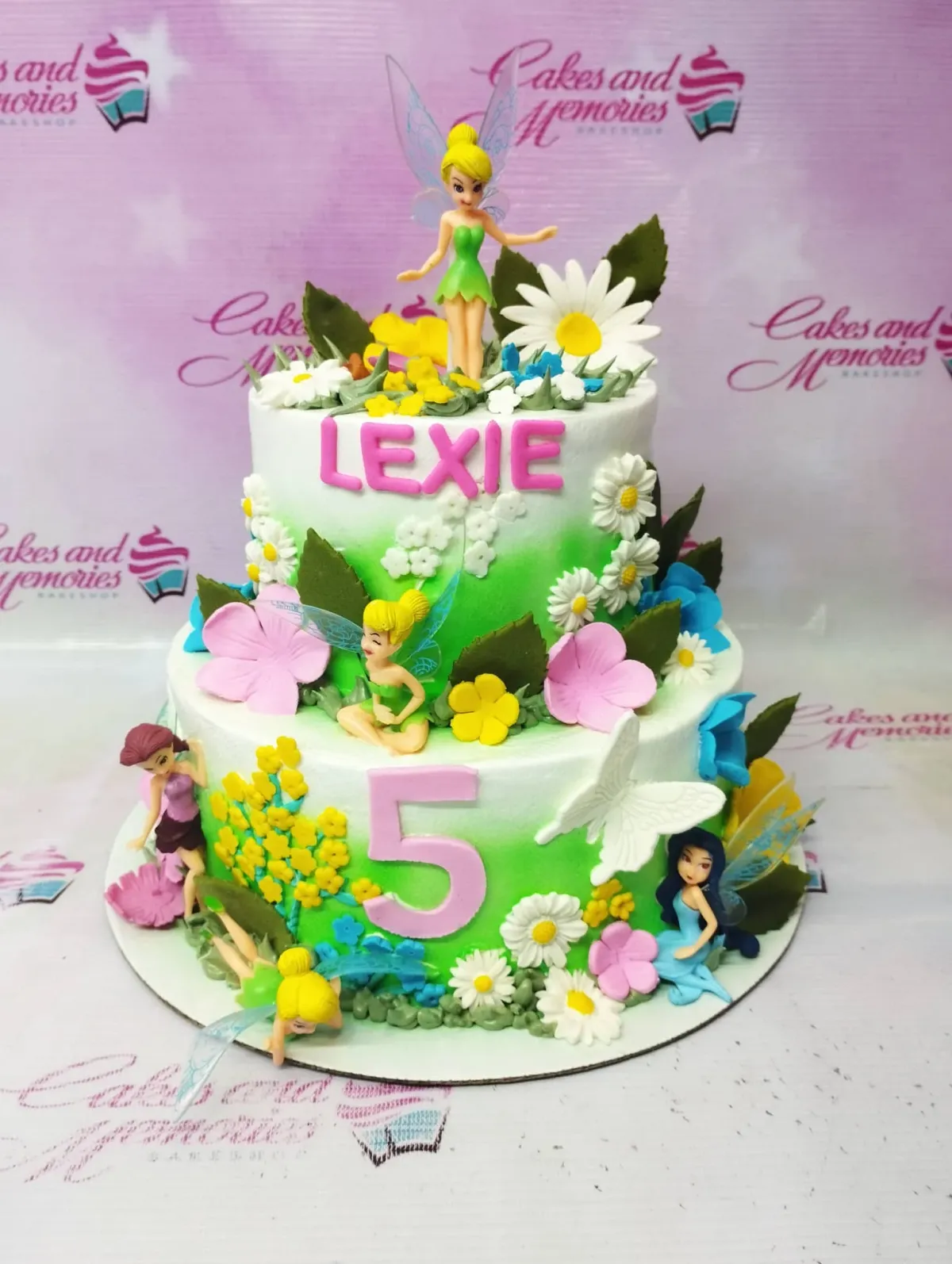 A green gradient and white two-tier cake themed as a Tinker Bell fairy garden, with fairy figurines, fondant flowers, and gumpaste letters. A green gradient and white two-tier cake themed as a Tinker Bell fairy garden, with fairy figurines, fondant flowers, and gumpaste letters.