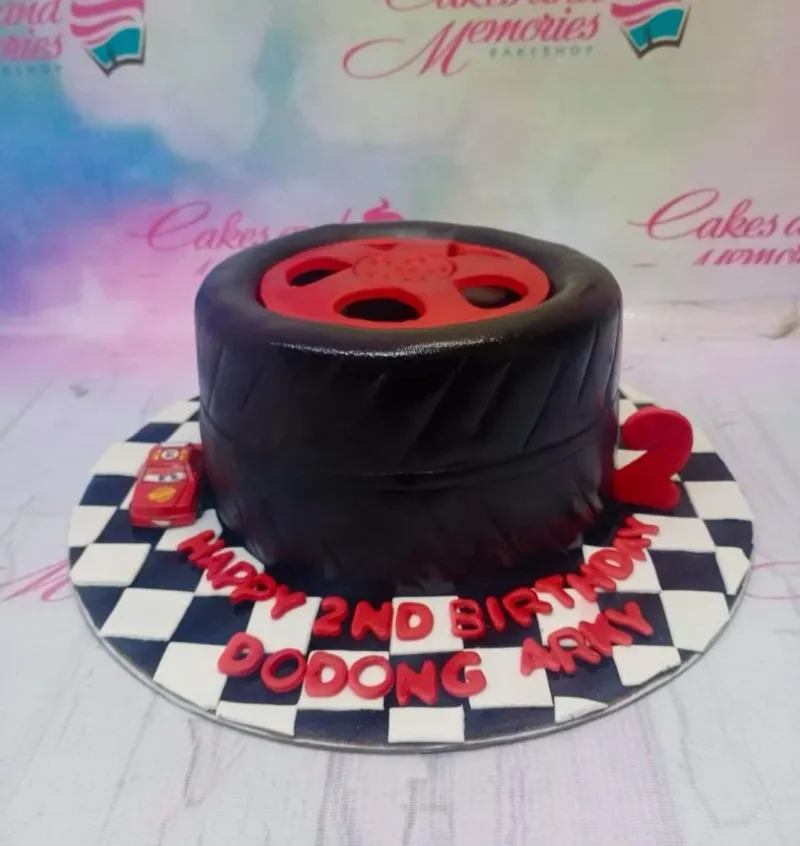 A black 1-tier fondant cake, tire-shaped with a red rim, a small red toy car, and a red number two, on a checkered base. A black 1-tier fondant cake, tire-shaped with a red rim, a small red toy car, and a red number two, on a checkered base.