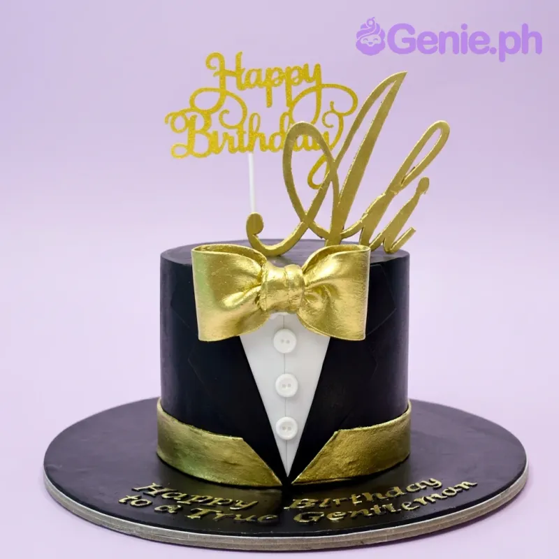 A black fondant cake, tuxedo-style, featuring a gold Happy Birthday Ali topper, bow tie, and white shirt buttons. A black fondant cake, tuxedo-style, featuring a gold Happy Birthday Ali topper, bow tie, and white shirt buttons.