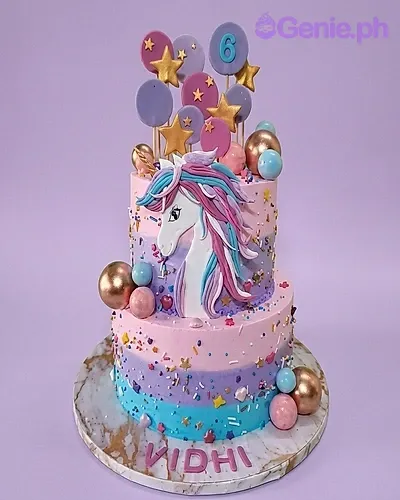 A pink, purple, and blue striped 2-tier cake themed around Unicorn, featuring a large unicorn face and a number six topper, with gold stars, colorful spheres, and 'VIDHI' text. A pink, purple, and blue striped 2-tier cake themed around Unicorn, featuring a large unicorn face and a number six topper, with gold stars, colorful spheres, and 'VIDHI' text.