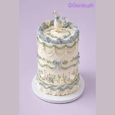 One tier cake with a duck figurine, white soft icing, blue icing swags, white and blue icing flowers, blue icing borders, blue icing bows, green icing leaves, white fondant flowers