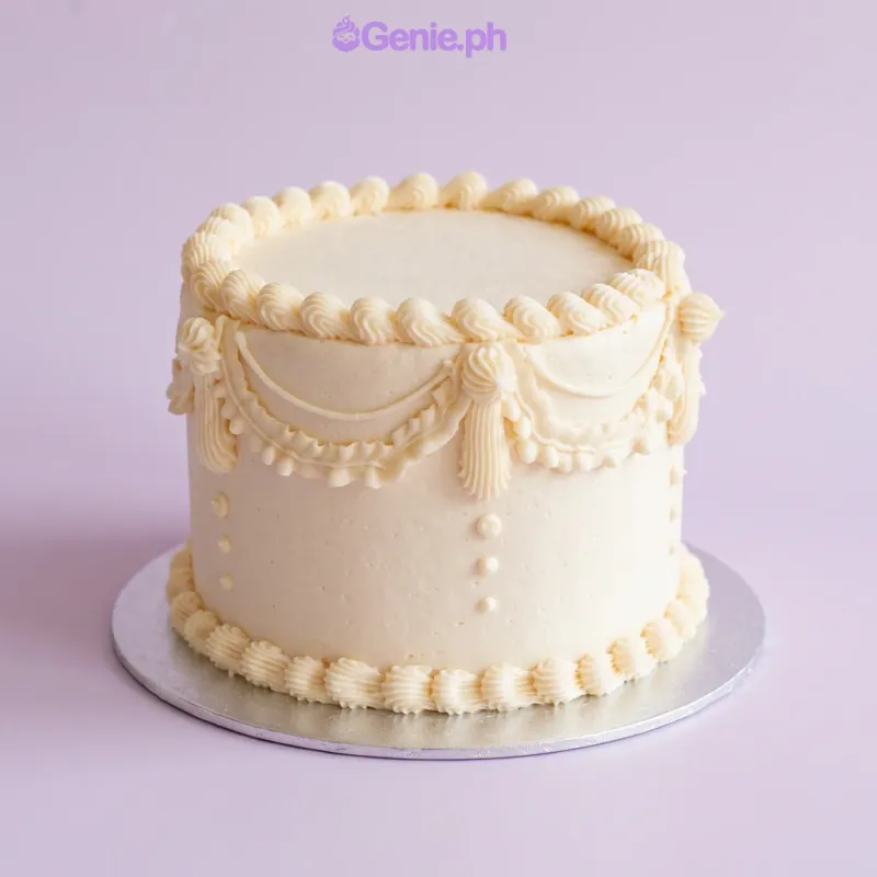Champagne-colored single-tier cake with soft icing, intricate piped swags, and delicate shell borders