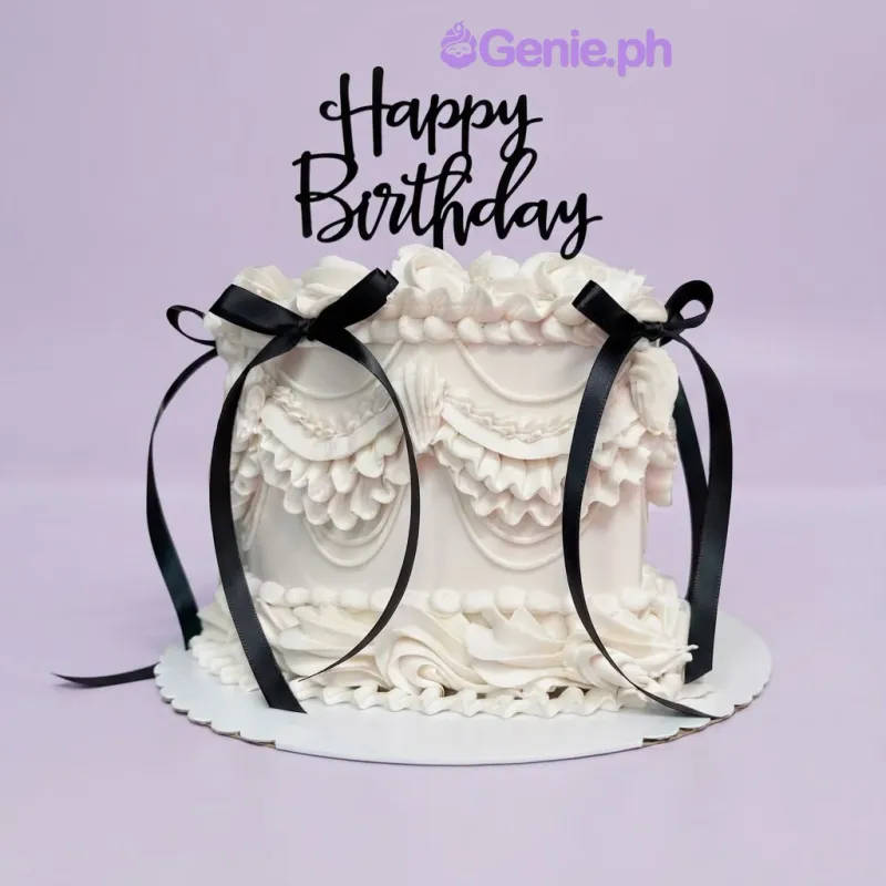 White 1-tier cake with soft icing, intricate piped swags and ruffles, black satin ribbons, and a black Happy Birthday topper