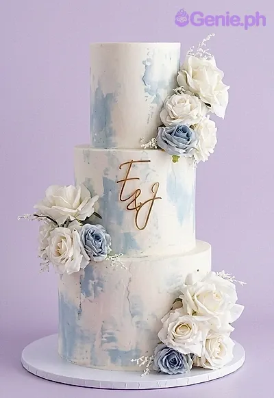 A white and blue three-tier cake themed around Watercolor Wedding, featuring a gold monogram topper and artificial white and light blue roses. A white and blue three-tier cake themed around Watercolor Wedding, featuring a gold monogram topper and artificial white and light blue roses.