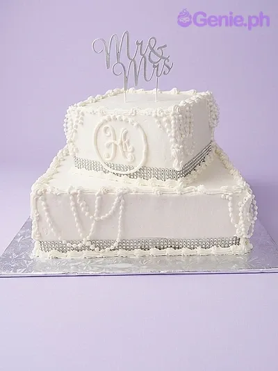 A white and silver 2-tier wedding cake, featuring an 'Mr & Mrs' script topper, piped monogram, silver dragees, and swags. A white and silver 2-tier wedding cake, featuring an 'Mr & Mrs' script topper, piped monogram, silver dragees, and swags.