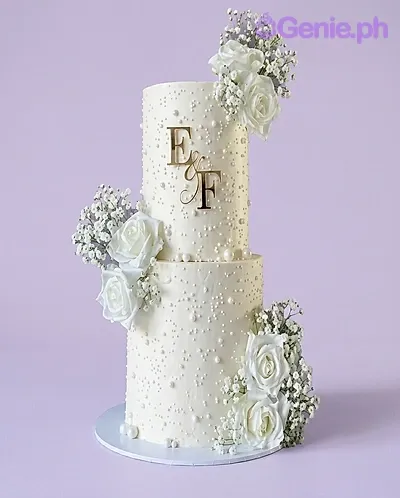 A white and gold 2-tier wedding cake, featuring gold initials 'E and F', white roses, baby's breath, and edible pearls. A white and gold 2-tier wedding cake, featuring gold initials 'E and F', white roses, baby's breath, and edible pearls.