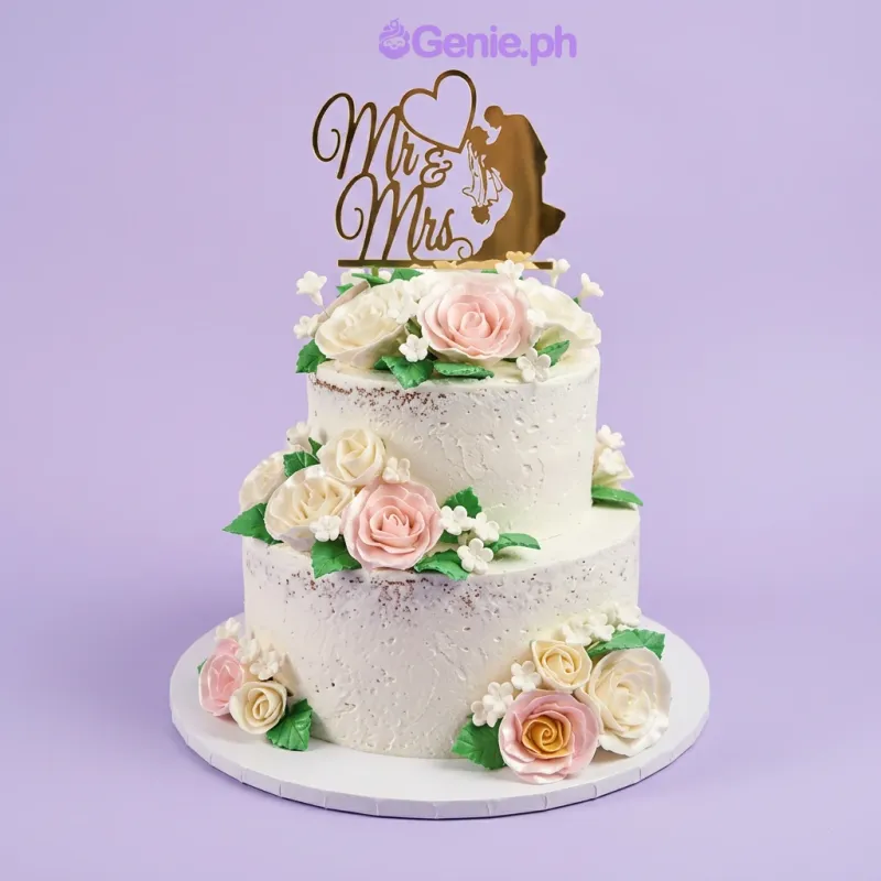 A white semi-naked two-tier wedding cake, featuring a gold Mr & Mrs topper, pink and white gumpaste roses, and green leaves. A white semi-naked two-tier wedding cake, featuring a gold Mr & Mrs topper, pink and white gumpaste roses, and green leaves.