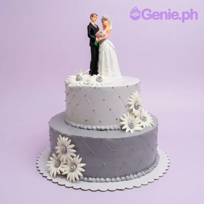 A gray and white 2-tier cake themed around Wedding, featuring a bride and groom figurine, white daisies, and a quilted pattern. A gray and white 2-tier cake themed around Wedding, featuring a bride and groom figurine, white daisies, and a quilted pattern.