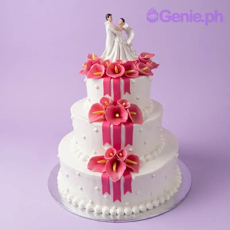 A white 3-tier wedding cake, adorned with pink calla lilies, red ribbons, white icing dots, and a bride and groom figurine topper. A white 3-tier wedding cake, adorned with pink calla lilies, red ribbons, white icing dots, and a bride and groom figurine topper.