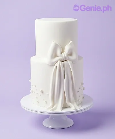 A white two-tier fondant cake themed around White Minimalist, featuring a large sculpted fondant bow and white pearl dragees. A white two-tier fondant cake themed around White Minimalist, featuring a large sculpted fondant bow and white pearl dragees.
