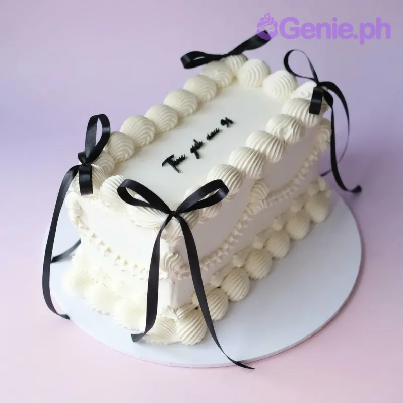 A white rectangular minimalist cake with black accents, featuring black satin ribbons and piped white borders, with a black message. A white rectangular minimalist cake with black accents, featuring black satin ribbons and piped white borders, with a black message.
