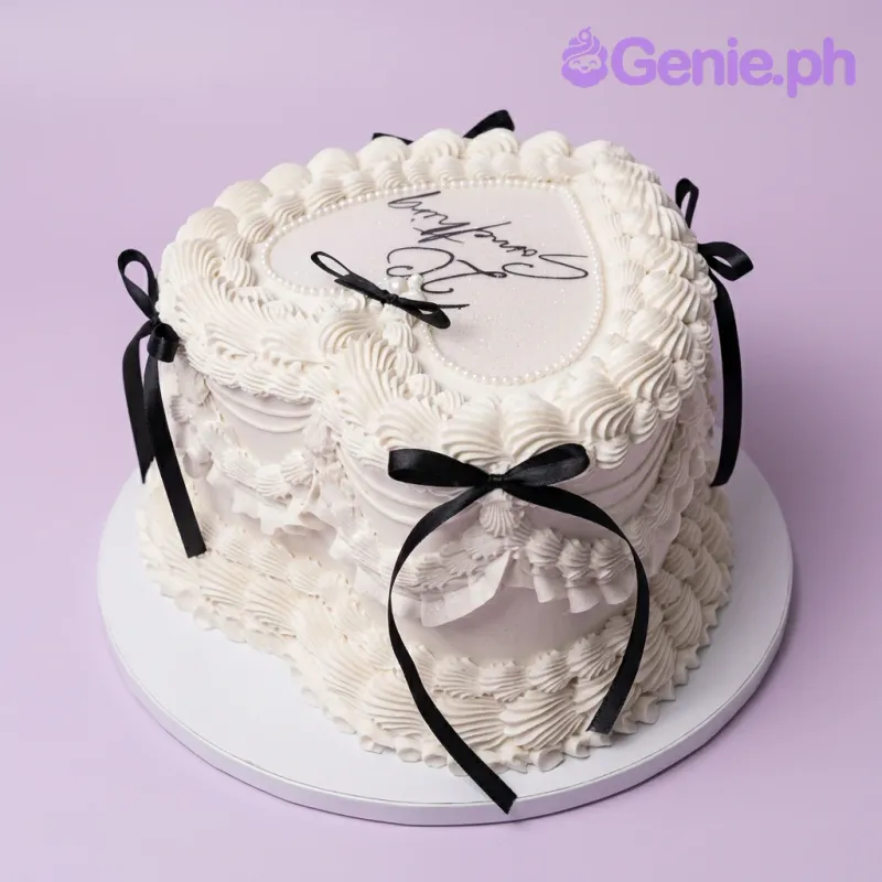 A white heart-shaped vintage 1-tier cake with piped ruffles, black satin bows, and '23 Something' text.