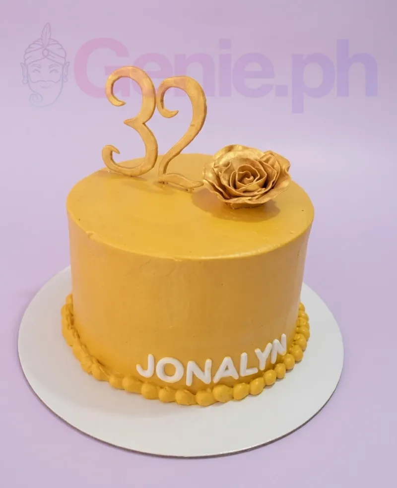 A yellow 1-tier cake with gold number 32 and rose toppers, white 'JONALYN' text, and a yellow beaded base border. A yellow 1-tier cake with gold number 32 and rose toppers, white 'JONALYN' text, and a yellow beaded base border.