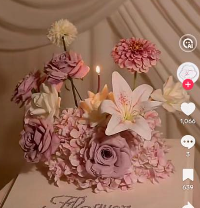 A cream 6-inch round cake featuring a lush arrangement of lavender roses, pink lilies, and small hydrangeas with a single lit candle.