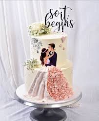 A photo of a So It Begins themed cake. It is a White 2 Tier cake decorated with a 'so it begins' text topper, a couple photo print, pink ruffled dress/skirt, white edible flowers, and silver leaf accents.
