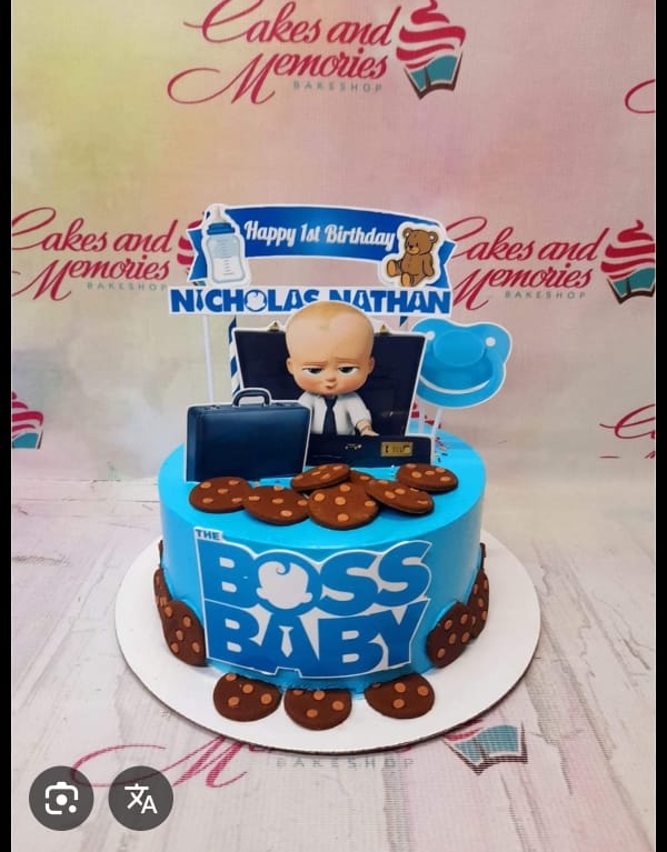 A photo of a Boss Baby themed cake. It is a custom colored 1 Tier Fondant cake decorated with a Boss Baby figure, a 'Happy 1st Birthday Nicholas Nathan' banner, a blue briefcase, a blue pacifier, chocolate chip cookies, and 'THE BOSS BABY' text on the side.