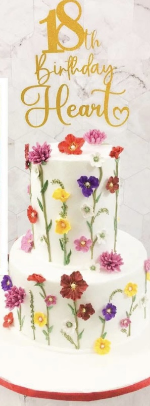 A photo of a Gelissa's 18th Birthday Floral themed cake. It is a white 2 Tier cake decorated with colorful edible flowers, green piped stems, and a gold glitter topper that says '18th Birthday Gelissa'.