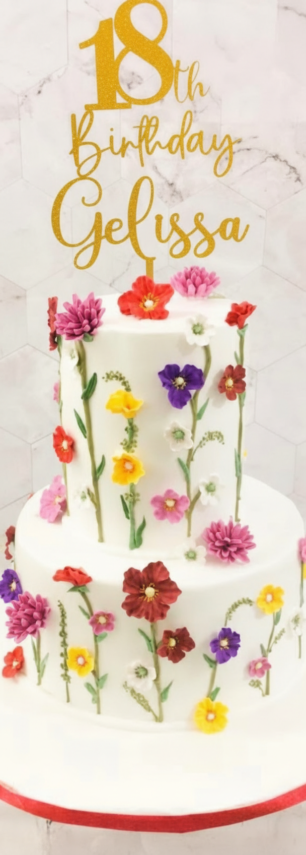 A photo of a Gelissa's 18th Birthday Floral themed cake. It is a white 2 Tier cake decorated with colorful edible flowers, green piped stems, and a gold glitter topper that says '18th Birthday Gelissa'.
