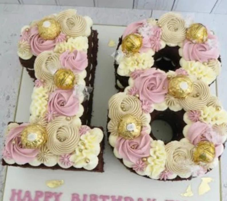 Number 18 shaped chocolate cake decorated with pink and cream icing rosettes, gold Ferrero Rocher chocolates, and gold sprinkles.