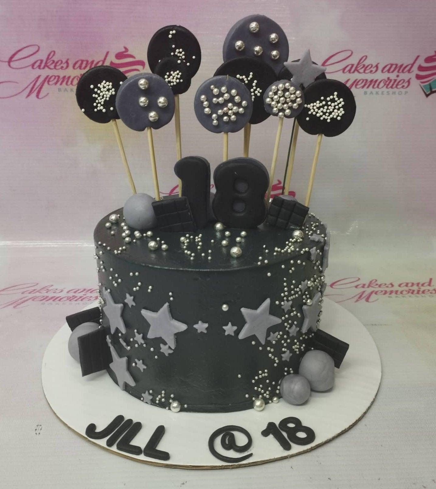 Black 18th birthday cake with silver and gray star decorations, chocolate bars, and round toppers with silver pearls.
