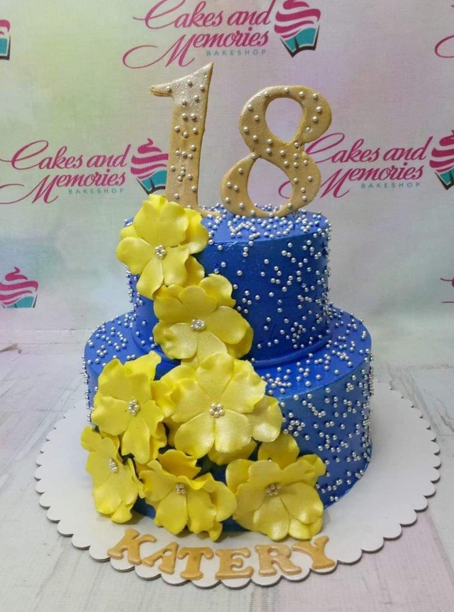 Two-tier royal blue cake with yellow gumpaste flowers, silver pearls, and a gold number 18 topper.